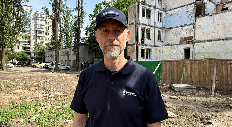 David Norlin i blå MCF-tröja framför resterna av ett bombat lägenhetshus.
