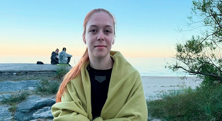 Porträtt på Polina Rahulina när hon sitter med havet och en rosa solnedgång i bakgrunden.