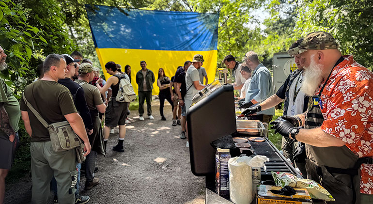 Deltagare i Repower står i kö till att få sig varsin hamburgare som serveras av frivilliga. Grillen stor utomhus i skogen och en stor ukrainsk flagga är uppspänd mellan träden.