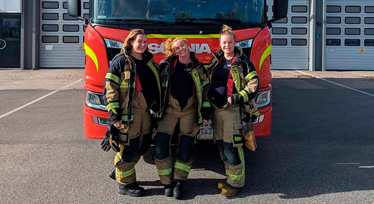 Tre gravida brandmän står framför en brandbil. Deras arbetsbyxor är uppknäppta i midjan på grund av gravidmagarna.