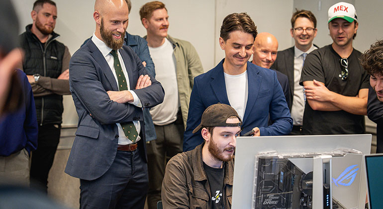 En man med bakåtvänd keps sitter framför en dataskärm. Bakom honom står flera personer, bland andra Carl-Oskar Bohlin och Faruk Pita.