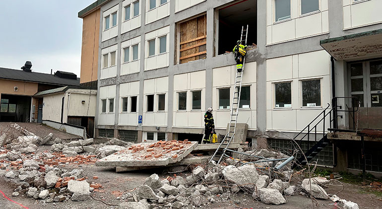 En före detta skola har utsatts för en fiktiv bomb, fönster är urblåsta och rasmassor täcker marken. En brandman är på väg upp för en stege för att ta sig in genom ett urblåst fönster, hans kollega står kvar på marken.