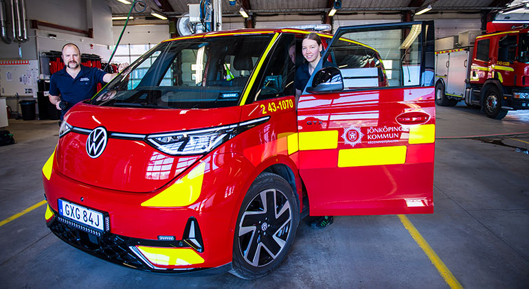 Elbrandbilen i ett garage, med två personer brandmän bredvid den.