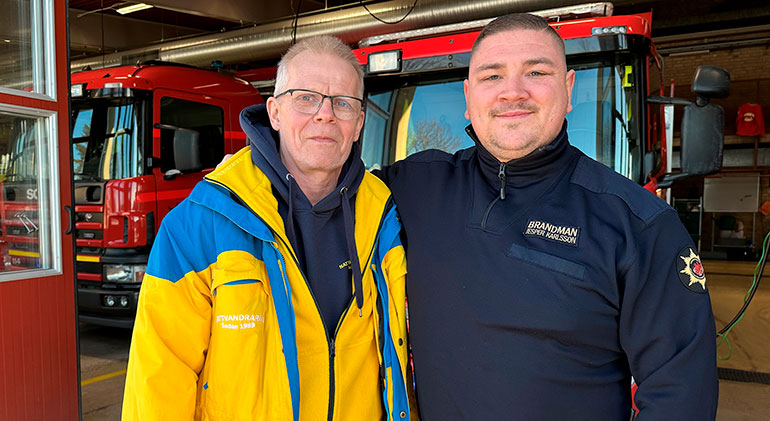 Stefan Nilsson, klädd i gul nattvandrarjacka och Jesper Karlsson, klädd i bli räddningstjänstkläder, håller om varandra. I bakgrunden ser man två brandbilar.